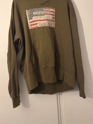 Olivgrön hoodie med amerikansk flagga - Olivgrön hoodie från Ralph Lauren med stor patch av amerikanska flaggan på bröstet. Hoodien har huva och känguruficka framtill. Tillverkad i mjukt bomullsmaterial och har en avslappnad passform. Perfekt för en casual och cool stil.