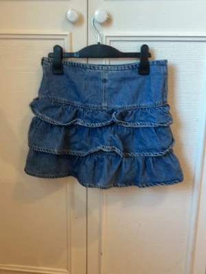 Blå jeanskjol med volanger - Supersöt blå jeanskjol med tre volanglager. Kjolen har en klassisk denimlook och är perfekt för att skapa en trendig och avslappnad stil. Passar till allt. Detta var en av mina favorit plagg men har tyvärr blivit för liten.