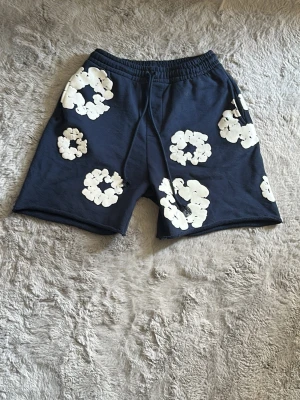 Mörkblå Denim tears shorts med vita tears - Snygga mörkblå shorts med stora vita blommor tryckta över hela plagget. Shortsen har resår i midjan och snörning för extra komfort. Perfekta för varma dagar och ger en chill vibe till din outfit.