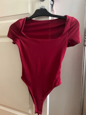 Vinröd body med kort ärm - Snygg vinröd body med korta ärmar och bred, fyrkantig urringning. Tillverkad i mjukt stretchigt material som sitter tight mot kroppen. Perfekt att styla med jeans eller kjol för en trendig look.
