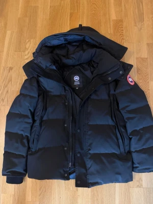 Svart dunjacka från Canada Goose - Säljer en svart dunjacka från Canada Goose med huva och klassisk logotyp på ärmen. Jackan har dragkedja och knappar framtill, två stora fickor och tjock vaddering som håller dig varm under kalla vinterdagar. Perfekt för dig som vill ha både stil och funktion den är i storlek M men sitter även lite stort på.