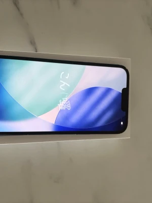  iPhone 14 Plus 256gb 85%hälsa  - A smartphone with a colorful screen displaying Japanese characters, placed on a white surface. The phone has a black frame and a notch at the top of the screen.