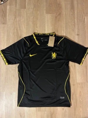 Svart Chelsea matchtröja 26/27  - Snygg svart Chelsea Matchtröja med gula detaljer och ett lejonmärke på bröstet. Tröjan har korta ärmar och gula sömmar samt mönster på krage och ärmslut. Tillverkad i ett lätt och ventilerande material, perfekt för träning eller match. HELT NY!