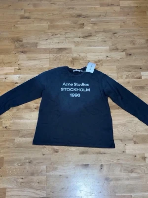 Svart långärmad topp Acne Studios - Svart långärmad topp från Acne Studios med tryck 'STOCKHOLM 1996' framtill. Klassisk rund halsringning och rak passform. Perfekt för dig som gillar stilrena och trendiga plagg.