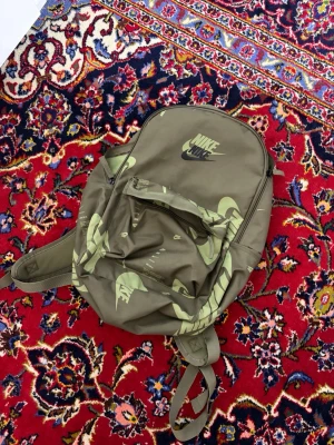 Olivgrön ryggsäck från Nike - Snygg olivgrön ryggsäck från Nike med stort logotryck i ljusgrönt och svart. Ryggsäcken har ett stort huvudfack, en mindre ytterficka med dragkedja och justerbara axelband. Perfekt för skola eller träning.