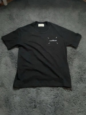 Svart t-shirt från Maison Margiela - Snygg svart t-shirt från Maison Margiela med diskret logga på bröstet. Klassisk rund halsringning och korta ärmar. Tillverkad i mjuk bomull för en skön känsla och enkel stil.