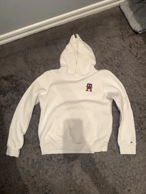 Vit hoodie från Tommy Hilfiger - Vit hoodie från Tommy Hilfiger med broderad TH-logga i rött och blått på bröstet. Klassisk känguruficka framtill och ribbade muddar. Huva med snygg randig detalj. Perfekt för en stilren och avslappnad look.