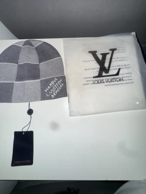 Gråvit Louis Vuitton mössa - Säljer en snygg gråvit mössa från Louis Vuitton med rutigt mönster och texten 'Marque Louis Vuitton Déposée' broderad på sidan. Mössan är stickad och har en klassisk passform. Perfekt accessoar för att lyfta din outfit.