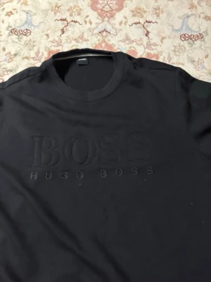 Svart sweatshirt från Hugo Boss - Säljer en svart sweatshirt från Hugo Boss med stor, upphöjd logga på bröstet. Tröjan har rund halsringning, långa ärmar och ribbade muddar vid ärmslut och nederkant. Perfekt för en clean och stilren look.