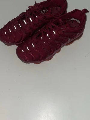 Nike VaporMax Plus. - Säljer ett par vinröda sneakers med cool transparent sula och reflekterande detaljer på ovandelen. Skorna har en unik design med vågiga mönster och snörning. Perfekta för dig som vill sticka ut med färg och stil.