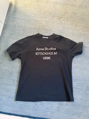 Svart Acne Studios t-shirt - Säljer en svart t-shirt från Acne Studios med tryck 'STOCKHOLM 1996' på bröstet. Klassisk passform och rund halsringning. Perfekt för dig som gillar stilrena och trendiga plagg med en touch av streetwear.