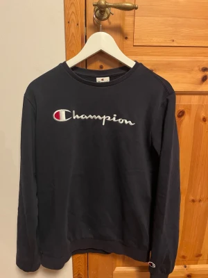 Svart sweatshirt från Champion 2XL - Snygg svart sweatshirt från Champion med det klassiska broderade logotypen i vitt och rött på bröstet. Tröjan har rund halsringning och långa ärmar, perfekt för en avslappnad streetwear-look.
