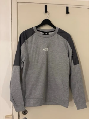 Grå sweatshirt från The North Face - Säljer en grå sweatshirt från The North Face med mörkgrå detaljer på axlar och ärmar. Tröjan har rund halsringning och logga på bröstet. Perfekt för en avslappnad och sportig stil. Materialet är mjukt och bekvämt.