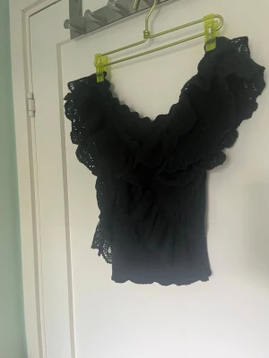 Svart volangblus med spetsdetaljer - Supersöt svart blus med djup v-ringning och stora volanger längs axlar och hals. Blusen är ärmlös och har fina spetsdetaljer som ger en romantisk känsla. Perfekt att styla med jeans eller kjol för en trendig look.