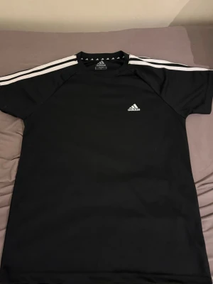 Svart Adidas Techfit t-shirt - Säljer en svart Adidas Techfit t-shirt med klassiska vita ränder på axlarna och Adidas-logga på bröstet. Tröjan är tillverkad i ett stretchigt funktionsmaterial som passar perfekt till träning och sportaktiviteter. Används inte längre har knappt använt den max 3 gånger men vill sälja den nu
