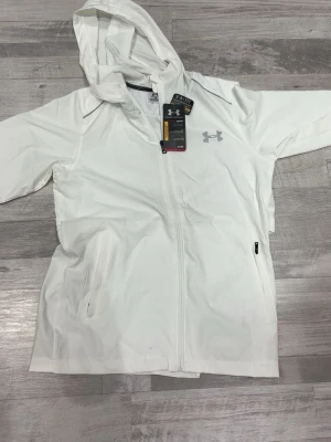 Vit vindjacka från Under Armour - Snygg vit vindjacka från Under Armour med huva och hel dragkedja framtill. Jackan har två sidofickor med dragkedja och logga på bröstet samt stor logga på ryggen. Tillverkad i lätt och vattenavvisande material, perfekt för blåsiga dagar.
