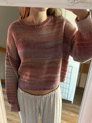 Stickad tröja i rosa toner - En croppad stickad tröja i mjuka rosa, röda och beige nyanser. Tröjan har rund hals, långa ärmar och ribbade muddar. Perfekt för en avslappnad och trendig stil. Materialet ser ut att vara mjukt och bekvämt.