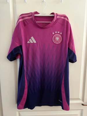 Tysklands fotbollströja rosa/lila - Snygg Tyskland fotbollströja från Adidas i rosa och lila med gradient mönster. Tröjan har korta ärmar, tre ränder på axlarna och Tysklands landslagsemblem med fyra stjärnor på bröstet. Tillverkad i lätt och ventilerande material, perfekt för träning eller match. (100% äkta köpt från adidas)