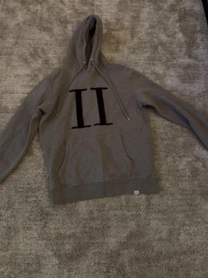 Les deux - Säljer en grå hoodie med stor svart II-logga på bröstet. Hoodien har huva med dragsko och en klassisk magficka framtill. Perfekt för dig som gillar enkel men snygg streetwear-stil.