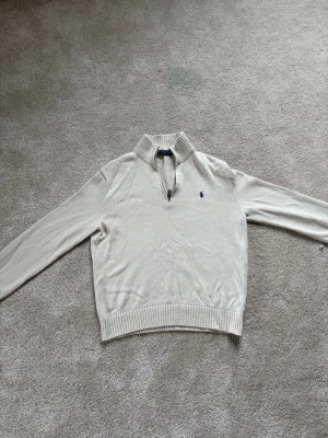 Beige stickad tröja med dragkedja - Snygg beige stickad tröja från Polo Ralph Lauren med liten blå logga på bröstet. Tröjan har en ribbad krage med kort dragkedja, långa ärmar och ribbade muddar vid ärmslut och nederkant. Perfekt för lager på lager och stilren look.