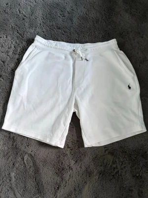 Vita shorts med snörning från Polo Ralph Lauren - Säljer ett par vita shorts från Polo Ralph Lauren med snörning i midjan och fickor på sidorna samt en bakficka. Klassisk logga broderad på ena benet. Perfekta för sommaren och tillverkade i mjuk bomull för maximal komfort.