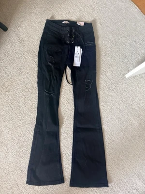 Svarta bootcut jeans med snörning - Säljer ett par svarta bootcut jeans med snörning i midjan och slitna detaljer på benen. Helt oanvända
