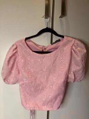 Rosa blus med puffärm - Supersöt rosa blus med broderade blommor och puffärmar. Toppen har snörning i ryggen och är croppad i modellen. Perfekt för sommaren. Använd 1 gång