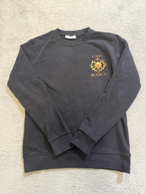 Svart sweatshirt från Casablanca - Snygg svart sweatshirt från Casablanca med tryck i guld och orange på bröstet där det står 'Casa Blanca' och ett coolt emblem. Tröjan har rund halsringning och långa ärmar. Perfekt för dig som gillar streetwear och vill ha något unikt i garderoben.