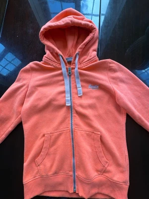 Korallfärgad hoodie med dragkedja - Säljer en korallfärgad hoodie med dragkedja och stora fickor framtill. Den har gråa snören i huvan och ribbade muddar vid ärmslut och nederkant. Superdry-logga broderad på bröstet. Perfekt för chill dagar eller när du vill vara bekväm.