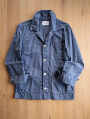 Blue Chore Jacket, size M - Snygg blå overshirt i bomull med klassisk krage och tre framfickor. Jackan har raka ärmar, vita knappar framtill och en avslappnad passform. Perfekt att slänga över en t-shirt för en casual look.