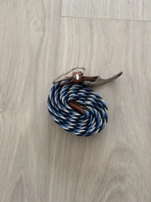 One wear bälte  - Säljer ett snyggt flätat bälte med randigt mönster i blått, vitt och brunt. Bältet har detaljer i brunt skinn och ett silverfärgat spänne. Perfekt för att ge din outfit en cool touch.