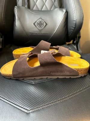 Mörk bruna Birkenstock sandaler i mocka - Säljer ett par klassiska Birkenstock sandaler i mörkbrunt mocka med två breda remmar och justerbara metallspännen. Fotbädden är gul och sulan har ett mönstrat grepp i brunt gummi. Perfekta för en avslappnad och stilren look.