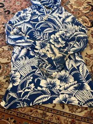 Blåvit hoodie från Polo Ralph Lauren - Snygg blå hoodie från Polo Ralph Lauren med tropiskt blommönster i vitt. Hoodien har huva med dragsko och är tillverkad i mjukt material. Perfekt för dig som vill sticka ut med en cool och unik stil.