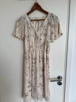 Blommig ljus klänning från H&M - Söt ljus klänning från H&M med blommigt mönster i beige och grå toner. Klänningen har v-ringning, korta ärmar och ett avslappnat fall. Perfekt för varma dagar och har en mjuk, luftig känsla.