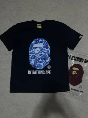 By bathing Ape - Bape T-shirt i svart med blå Bape-logo. Size S. Snygg och stilren streetwear T-shirt med en clean design och en riktigt nice färgkombination. Skön och bekväm att bära, perfekt till både vardags och streetwear outfits.
