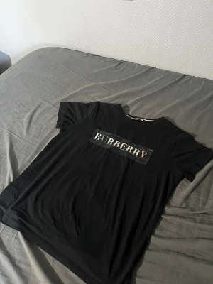 Svart Burberry t-shirt med logga - Snygg svart t-shirt från Burberry med klassisk logga framtill i vitt och rött. T-shirten har rund hals och korta ärmar, tillverkad i mjuk bomull för skön känsla. Perfekt för dig som vill ha en stilren och trendig look.