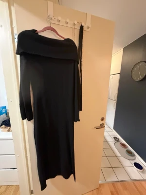 Svart lång stickad offshoulderklänning - Snygg svart långklänning i stickat material med offshoulder-design och lång ärm. Klänningen har en slits på sidan för extra stil och rörelsefrihet. Perfekt för dig som vill ha en elegant och bekväm look.
