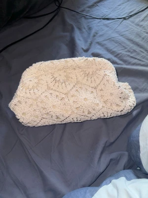 Beige virkad kuvertväska - Säljer en beige kuvertväska med virkat spetsmönster. Väskan har en mjuk och flexibel form och stängs upptill. Perfekt för dig som gillar en bohemisk och unik stil. Passar till många olika outfits och är lätt att bära med sig.