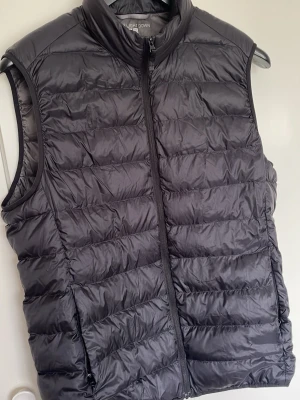 Uniqlo - Snygg grå dunväst med quiltad design och hög krage. Västen har en rak passform och stängs med dragkedja framtill. Perfekt att slänga över en hoodie eller tröja för extra värme. Materialet är lätt och smidigt, vilket gör den enkel att bära och styla.