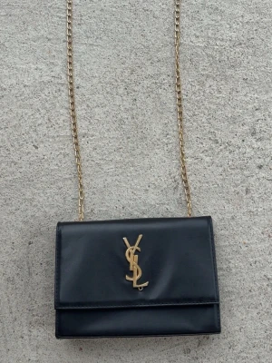Svart axelväska från Saint Laurent - Snygg svart axelväska i slätt läder med guldfärgad kedjerem och ikonisk YSL-logga framtill. Väskan har en stilren, rektangulär form och passar perfekt för dig som gillar klassisk men trendig design. Perfekt accessoar för att lyfta vilken outfit som helst.