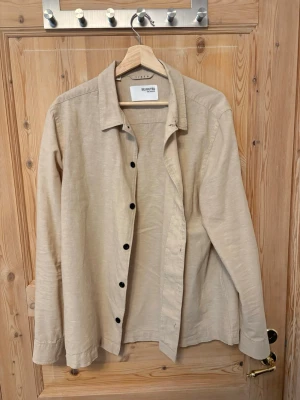 Beige linneskjorta från Selected Homme - Stilren beige skjorta i linne från Selected Homme. Skjortan har klassisk krage, svarta knappar och lång ärm. Perfekt för en avslappnad men snygg look. Passar bra till jeans eller chinos.