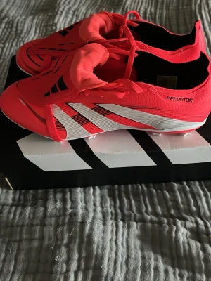 Adidas Predator League fotbollsskor - Säljer ett par Adidas Predator League FT FG/MG fotbollsskor i röd och vit färg med svarta detaljer. Skorna har snörning, låg profil och är tillverkade i syntetmaterial. Perfekta för spel på både fasta och mixade underlag. Skriv vid funderingar!