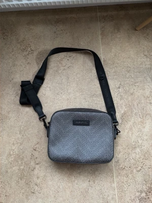 Grå axelväska från Valentino - Snygg grå axelväska från Valentino med justerbar svart axelrem. Väskan har ett diskret mönster med Valentino-loggan över hela ytan, dragkedja upptill och en extra ficka med dragkedja på baksidan. Perfekt storlek för det viktigaste och stilren design.