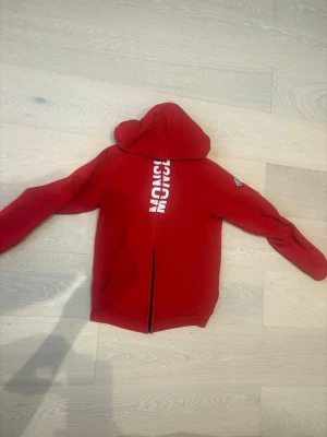 Röd hoodie från Moncler med tryck - Snygg röd hoodie från Moncler med vit logotyp-tryck på bröstet och klassisk Moncler-patch på ärmen. Hoodien har huva och dragkedja framtill. Perfekt för dig som vill ha en streetwear-look med premiumkänsla.