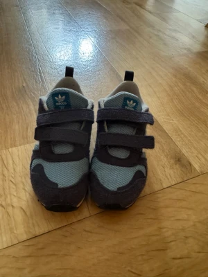 Blå sneakers från Adidas med kardborre - Snygga blå sneakers från Adidas med klassiska vita ränder på sidan och praktiska kardborreband. Ovandelen är i mesh och mockaliknande material, vilket ger både stil och komfort. Perfekta för sport eller vardagsaktiviteter.