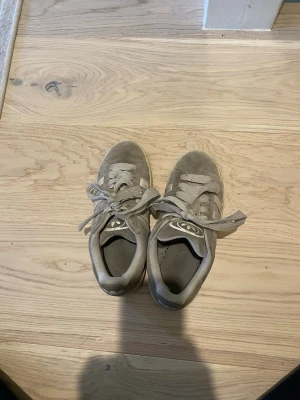 Grå sneakers i mocka från Adidas - Säljer ett par grå sneakers från Adidas i mocka med klassiska tre ränder på sidan och Adidas-logga på plösen. Skorna har breda snören och en låg profil, perfekta för en avslappnad stil.