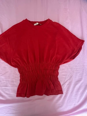 Röd topp med smockad midja - Snygg röd topp med rund halsringning och smockad midja som ger en markerad siluett. Toppen har korta ärmar och lätt volang nedtill. Perfekt att styla med jeans eller kjol för en trendig look.
