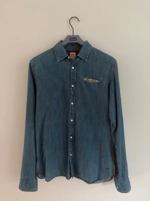Blå jeansskjorta med blommigt innerfoder bröstficka - Snygg blå jeansskjorta med långa ärmar och vita knappar. 100% bomull. Bröstfickan har ett blommigt innerfoder som sticker ut och ger en unik detalj. Skjortan har klassisk krage och avslappnad passform, perfekt för en cool och avslappnad stil. Vintage inspirerad, passar perfekt för lite retrolook. Mycket bra skick. Naturlig färgskiftning/slitning som ska se ut så.