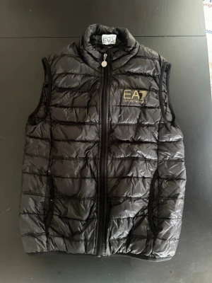 Svart dunväst från EA7 Emporio Armani - Snygg svart dunväst från EA7 Emporio Armani med guldfärgad logga på bröstet. Västen har hög krage, dragkedja framtill och två sidofickor. Quiltad design med lätt glansigt material som ger en modern look.      Snabb leverans 📦 