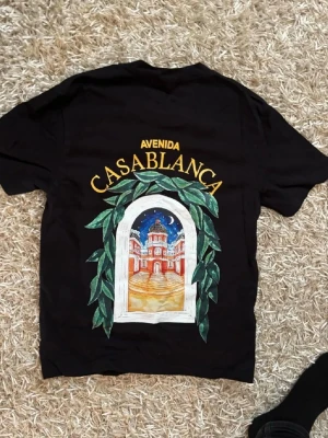 Casablanca tshirt  - Svart t-shirt från Casablanca med färgglatt tryck på ryggen som föreställer en byggnad genom ett fönster med gröna blad runtom och texten 'Avenida Casablanca'. Mindre tryck på bröstet. Klassisk passform och rund hals.